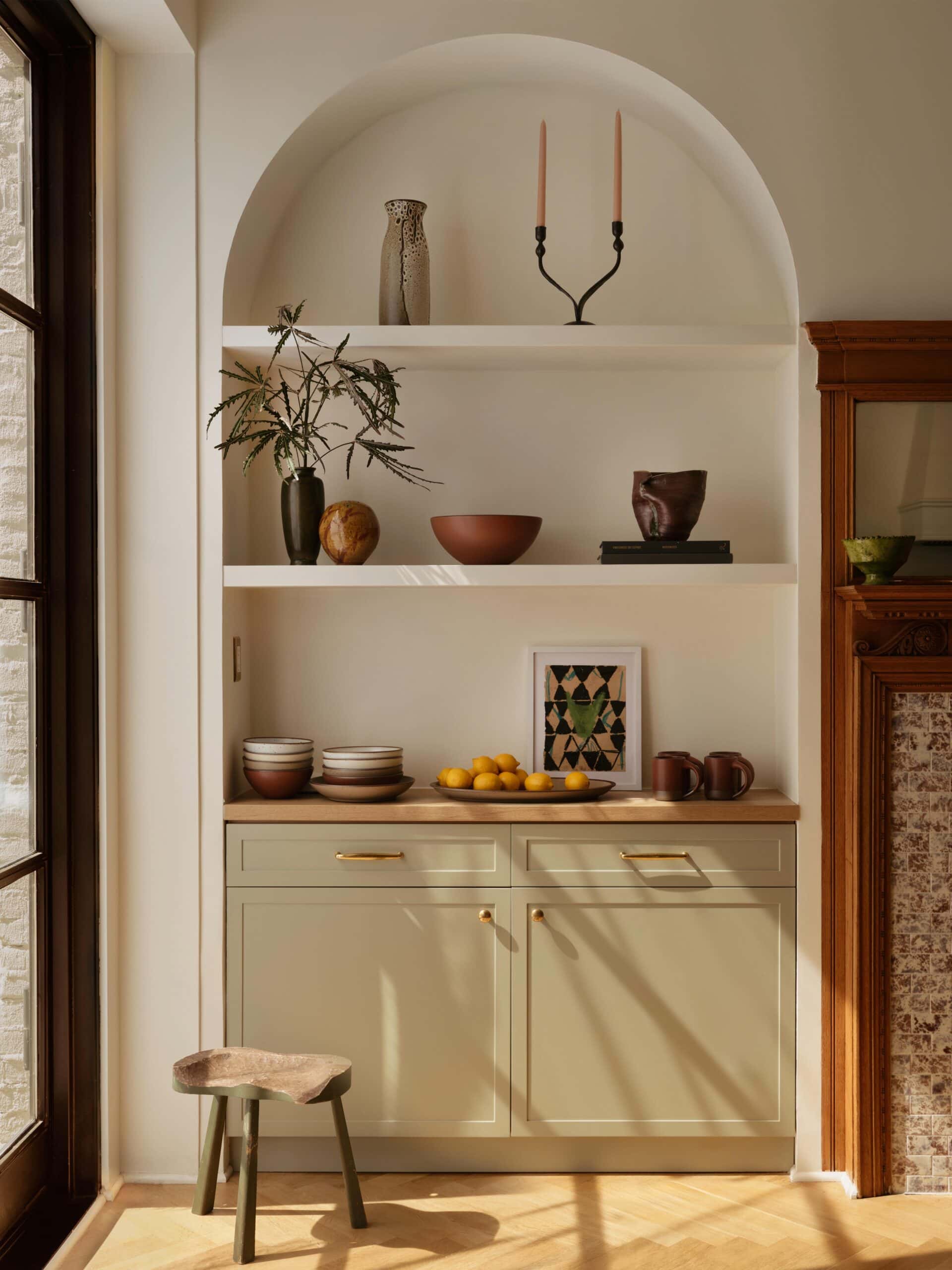 A built-in shelving unit with decorative objects, bowls, cups, a framed print, and a cluster of lemons, next to a window and wooden fireplace mantel.