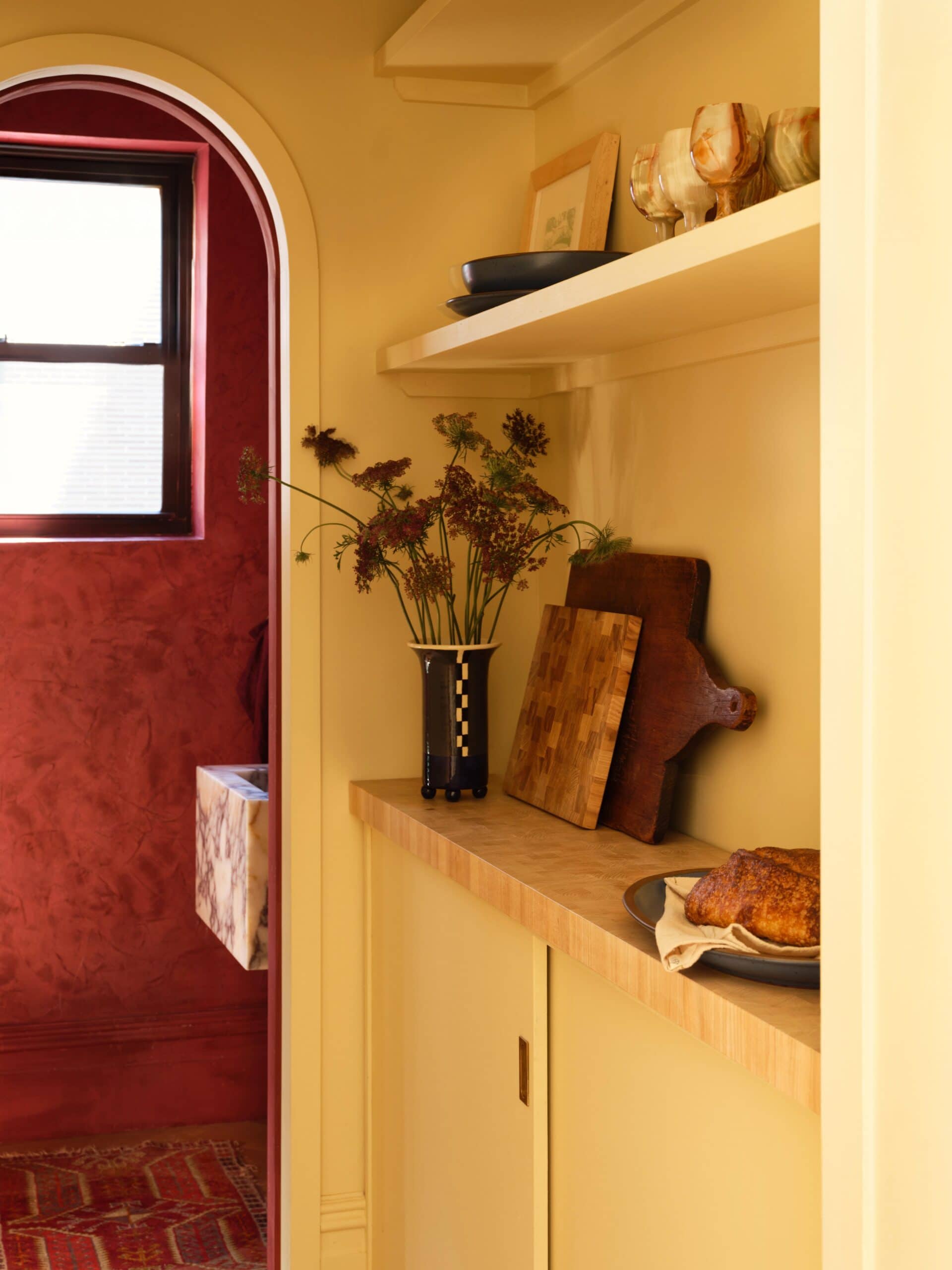 A cozy kitchen corner with yellow cabinets, wooden shelves holding art and glassware, a vase with flowers, cutting boards, and a loaf of bread; a red-walled room visible through an arched doorway.