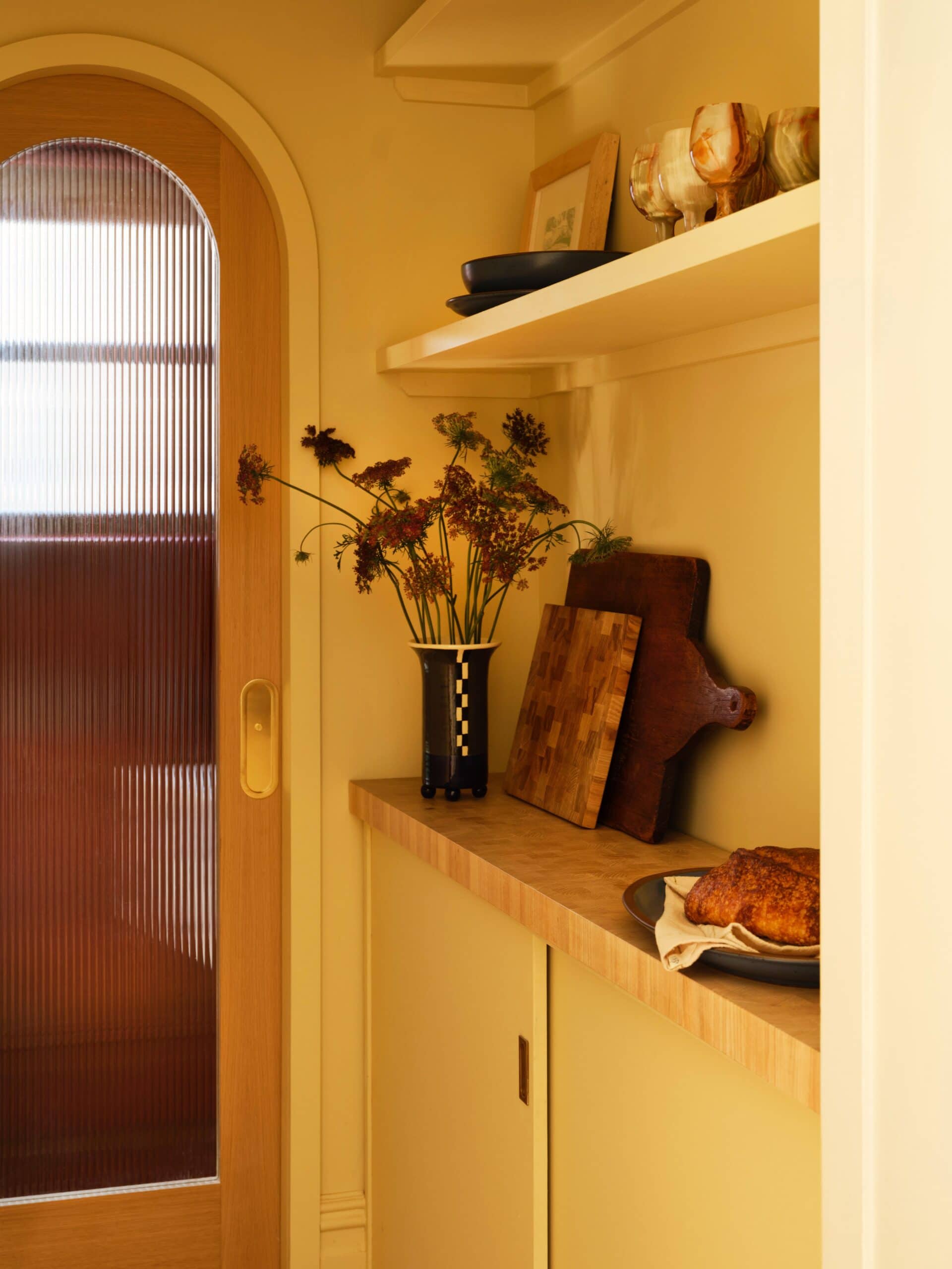 A narrow kitchen space with wooden shelves holding dishes, a vase of flowers, cutting boards, and a loaf of bread; a glass-paned door lets in natural light.