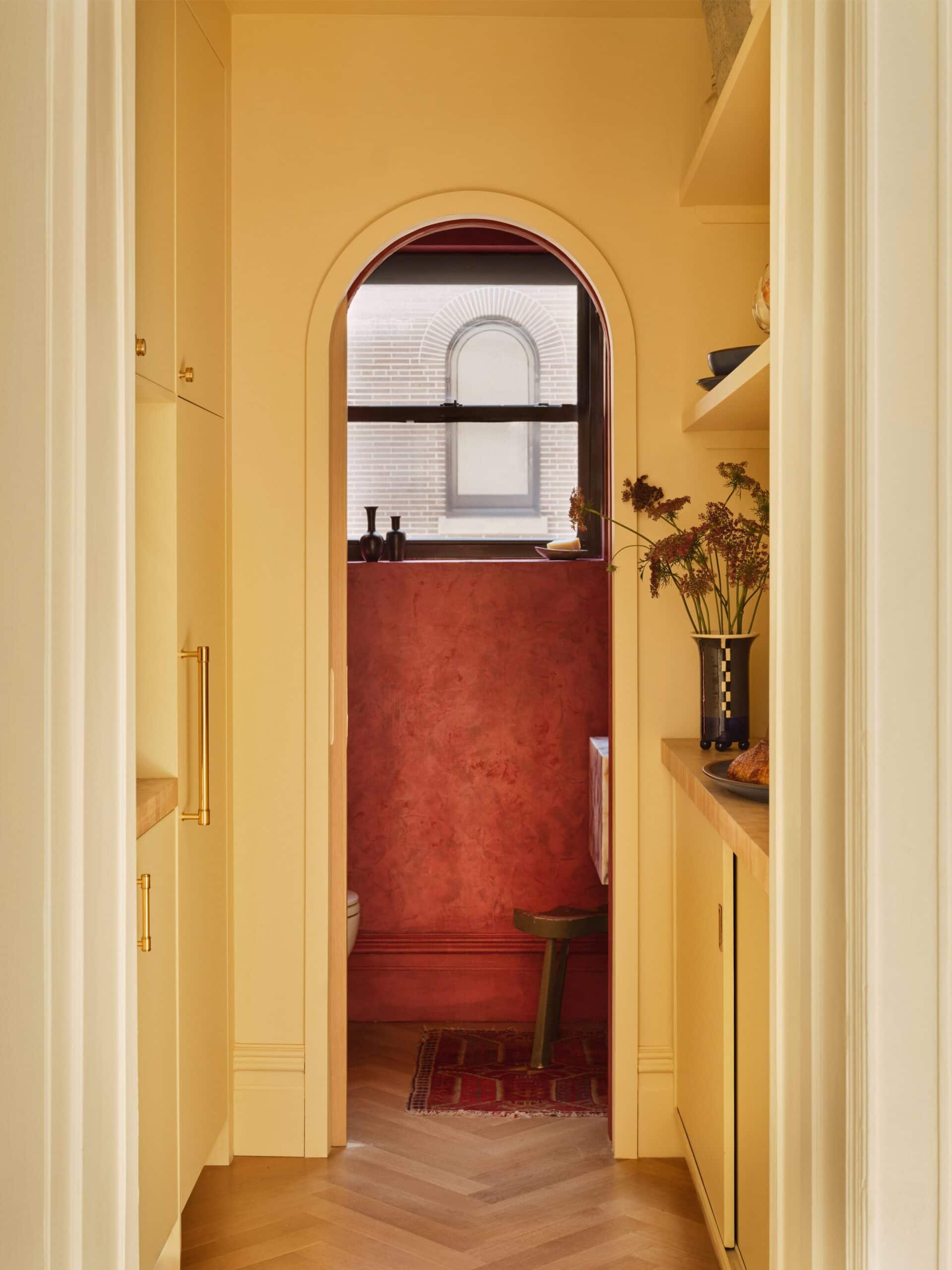 A small, warm-toned hallway with built-in cabinets leads to an arched doorway revealing a red-walled room, stool, window, and decorative items on a shelf.