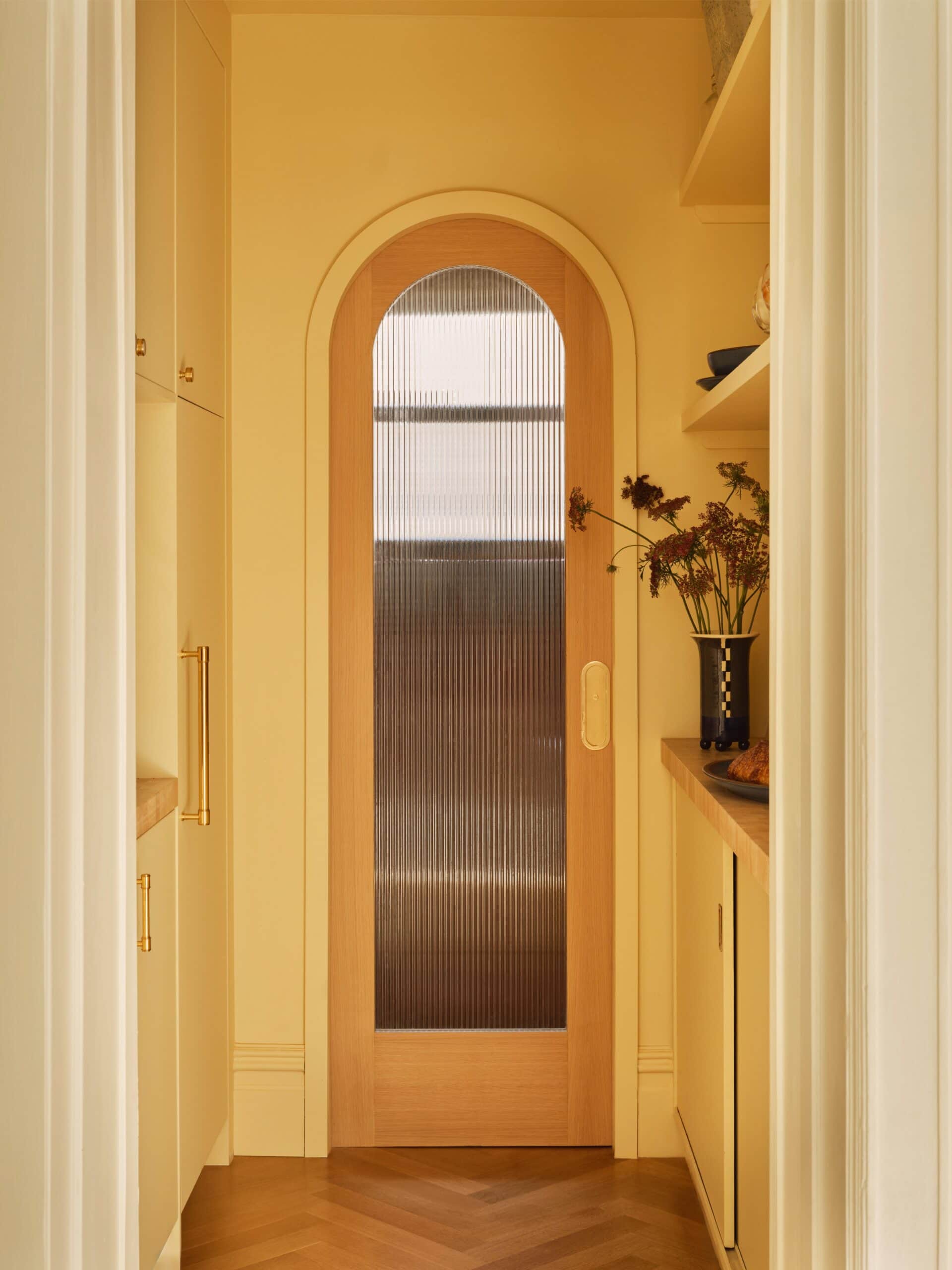 A hallway features a wooden door with an arched top and ribbed glass panels, surrounded by yellow cabinetry and shelves with dried flowers in a vase.