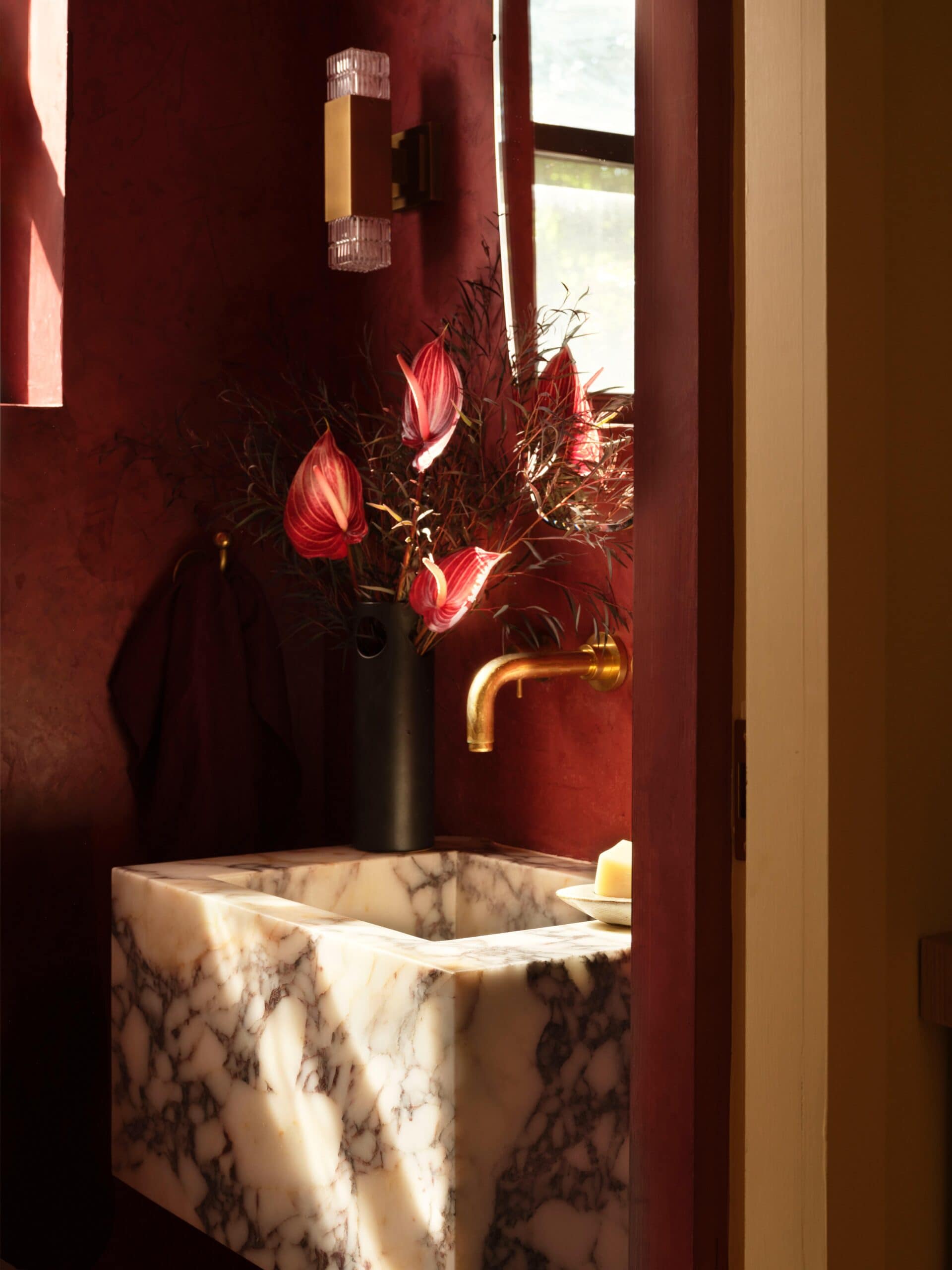 A marble sink with brass faucet, a black vase with pink flowers, a wall sconce, and a towel hanging on a dark red wall in a sunlit bathroom.