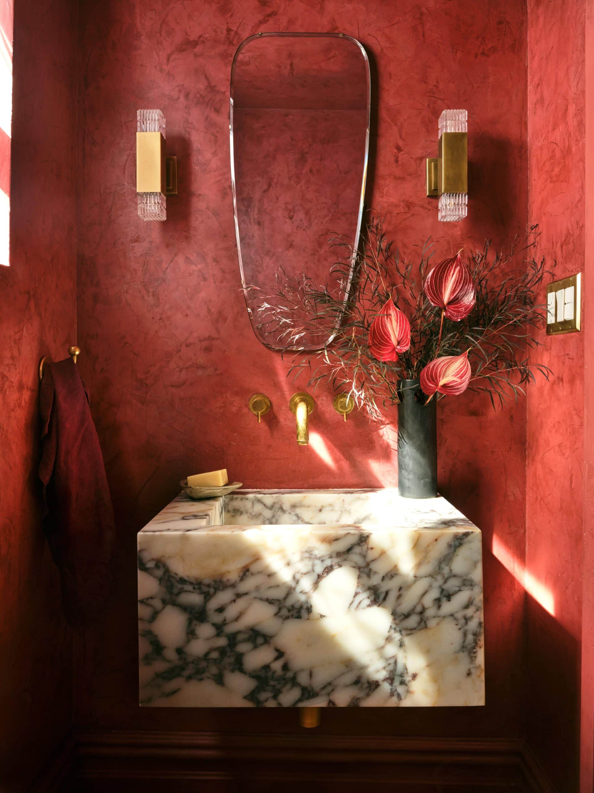 A marble sink with gold fixtures is set against red textured walls, beneath a curved mirror. Two wall sconces and a black vase with flowers complete the decor.