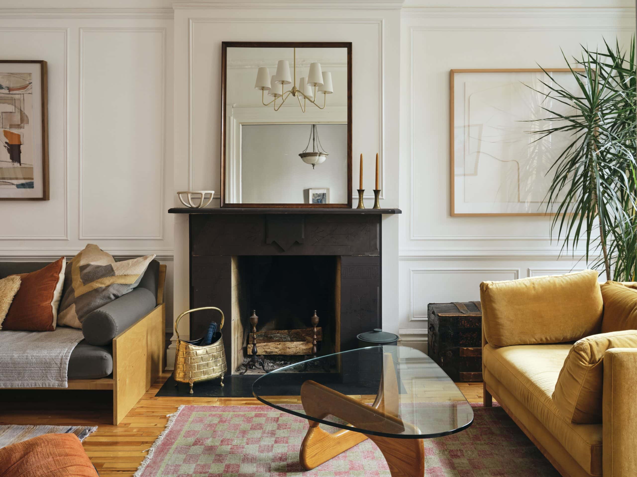 A living room with a black fireplace, a glass coffee table, a yellow sofa, a gray sofa, wall art, a large plant, and wooden floors.