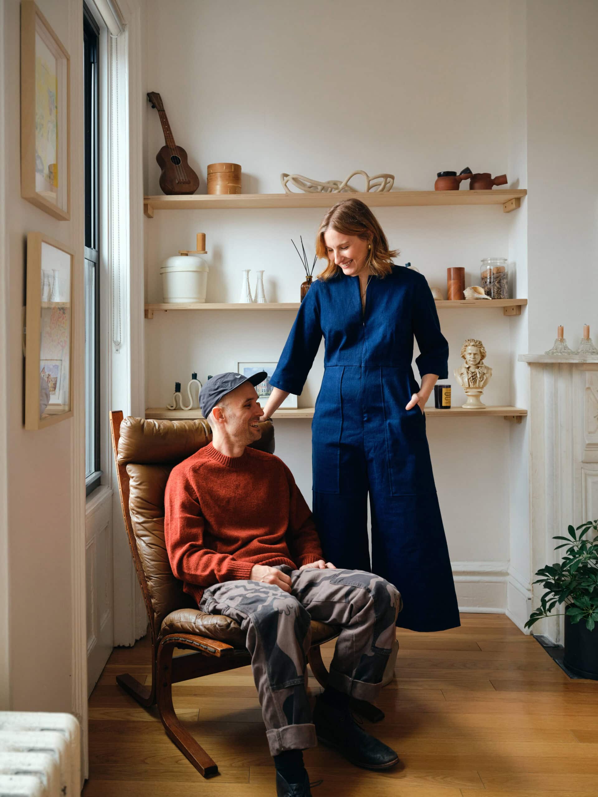 A seated man in a red sweater and cap looks up at a standing woman in a blue jumpsuit; they are in a cozy, well-lit room with shelves and decor.