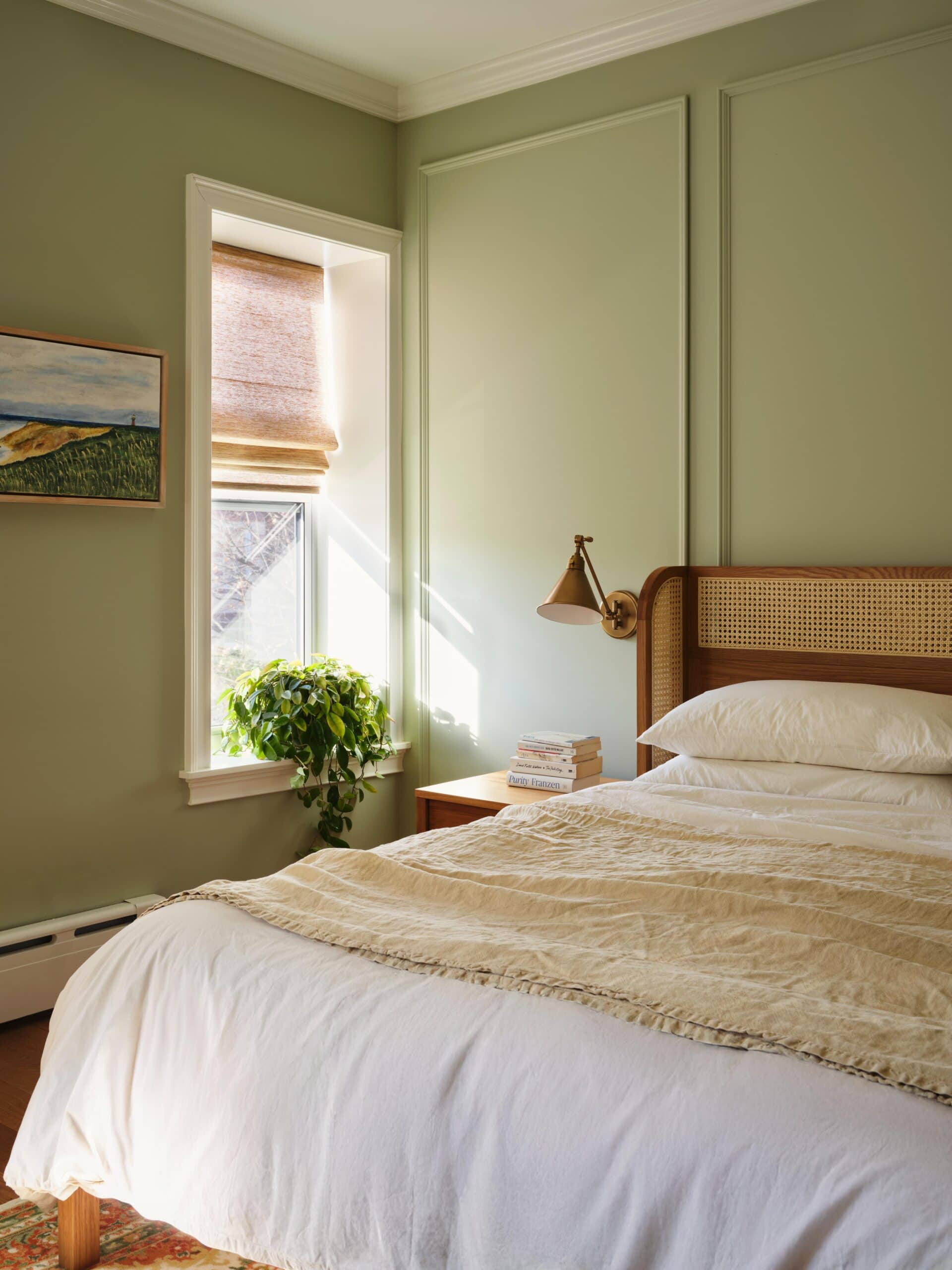 A neatly made bed with white bedding sits beside a window with a Roman shade, a potted plant, and a stack of books on a nightstand in a green-painted bedroom.