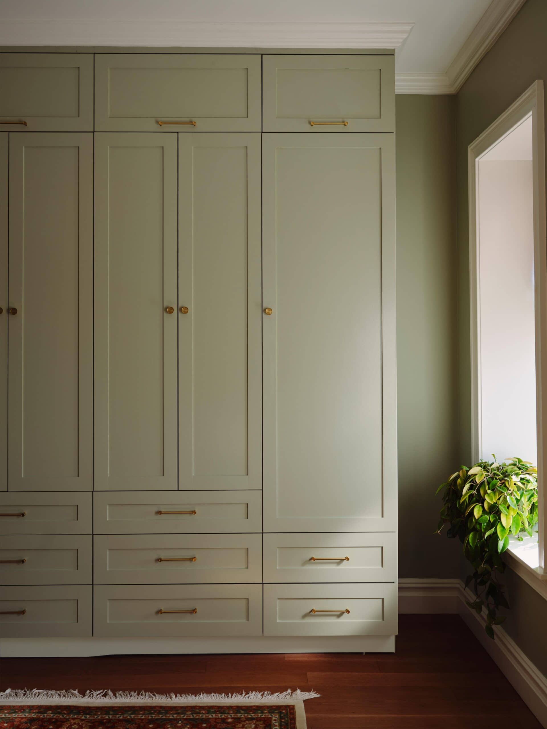 Tall, sage green built-in wardrobe with gold handles and knobs, multiple drawers and cabinets, next to a window with a potted plant on the floor.