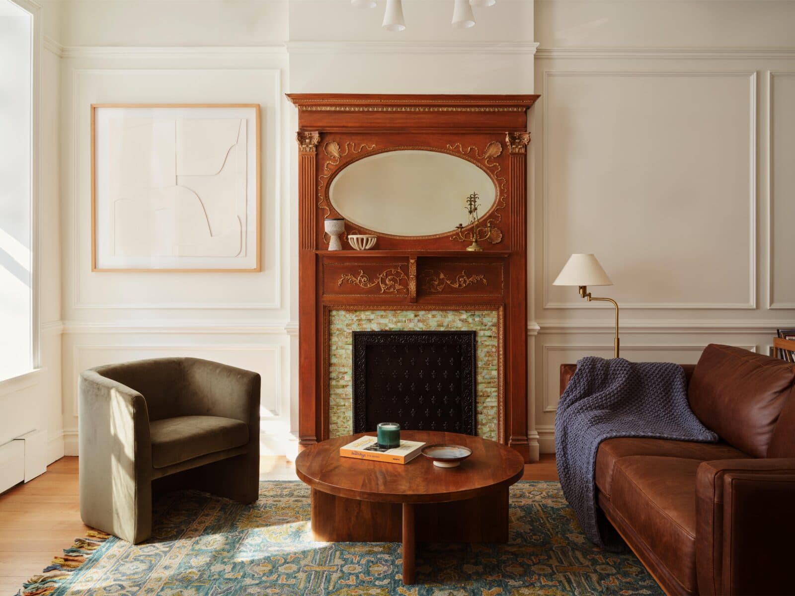 A living room with a brown leather sofa, green armchair, round wooden coffee table, and an ornate wooden fireplace with a mirror above it. A floor lamp and framed art are also present.