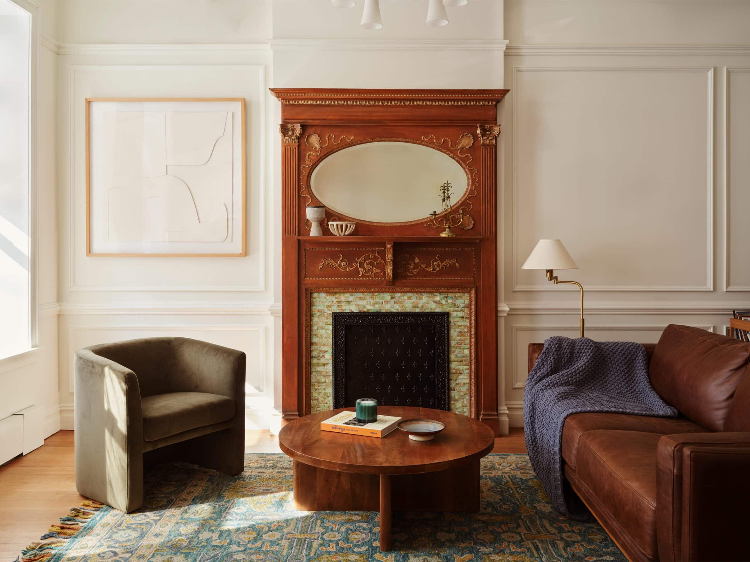 A living room with a brown leather sofa, green armchair, round wooden coffee table, and an ornate wooden fireplace with a mirror above it. A floor lamp and framed art are also present.