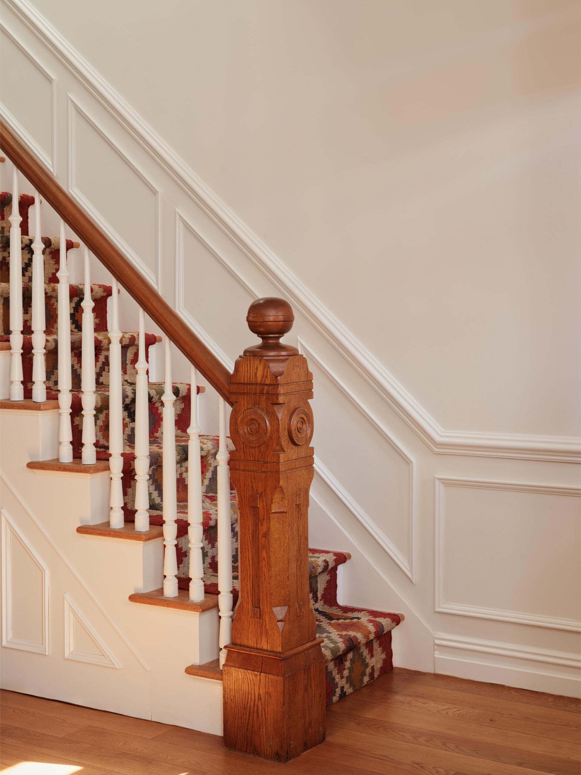 Wooden staircase with a patterned carpet runner, white balusters, and a decorative newel post, set against a wall with white wainscoting.
