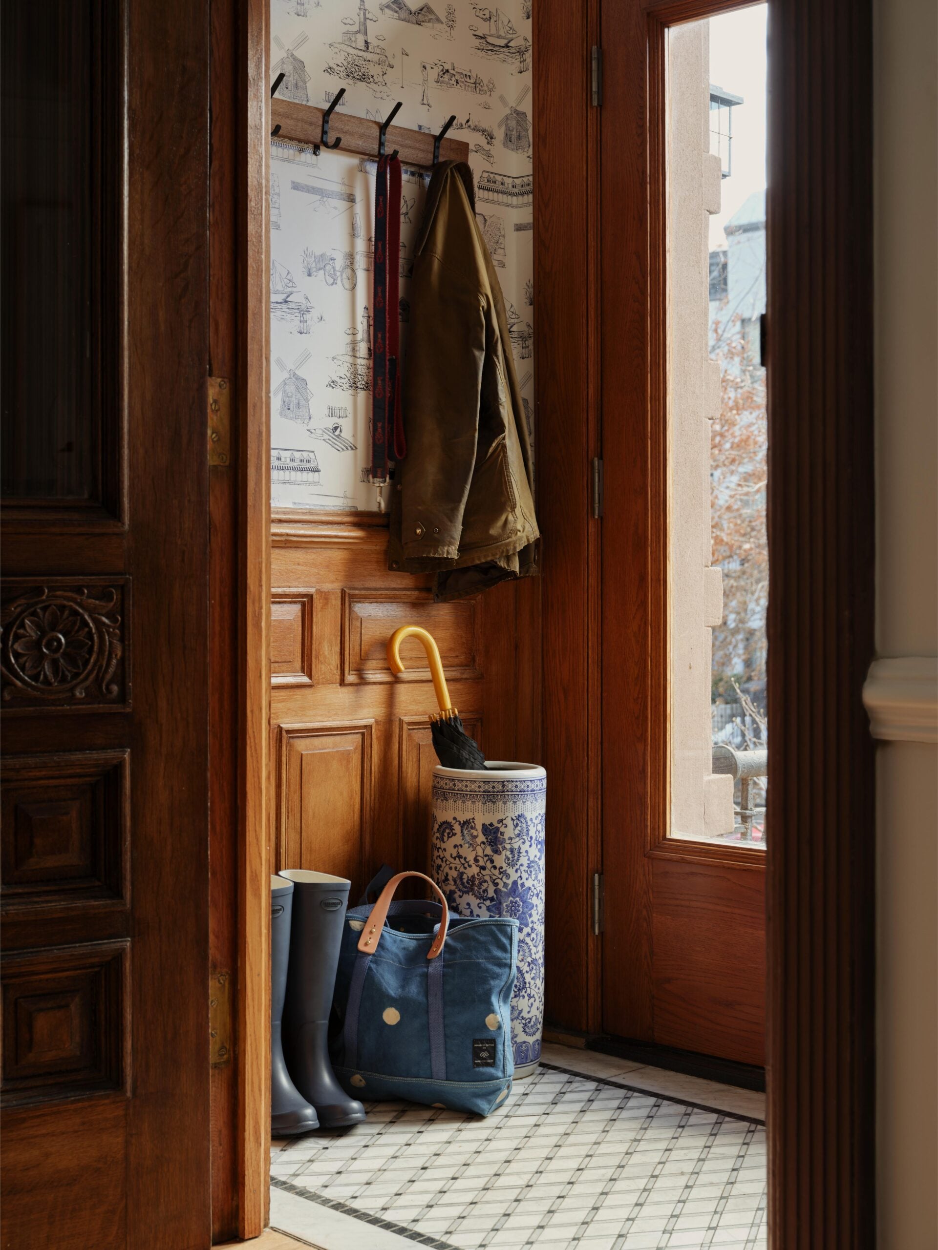 A small entryway with wooden walls, a coat and dog leash on hooks, rain boots, a blue bag, an umbrella in a ceramic stand, and a glass door showing trees outside.