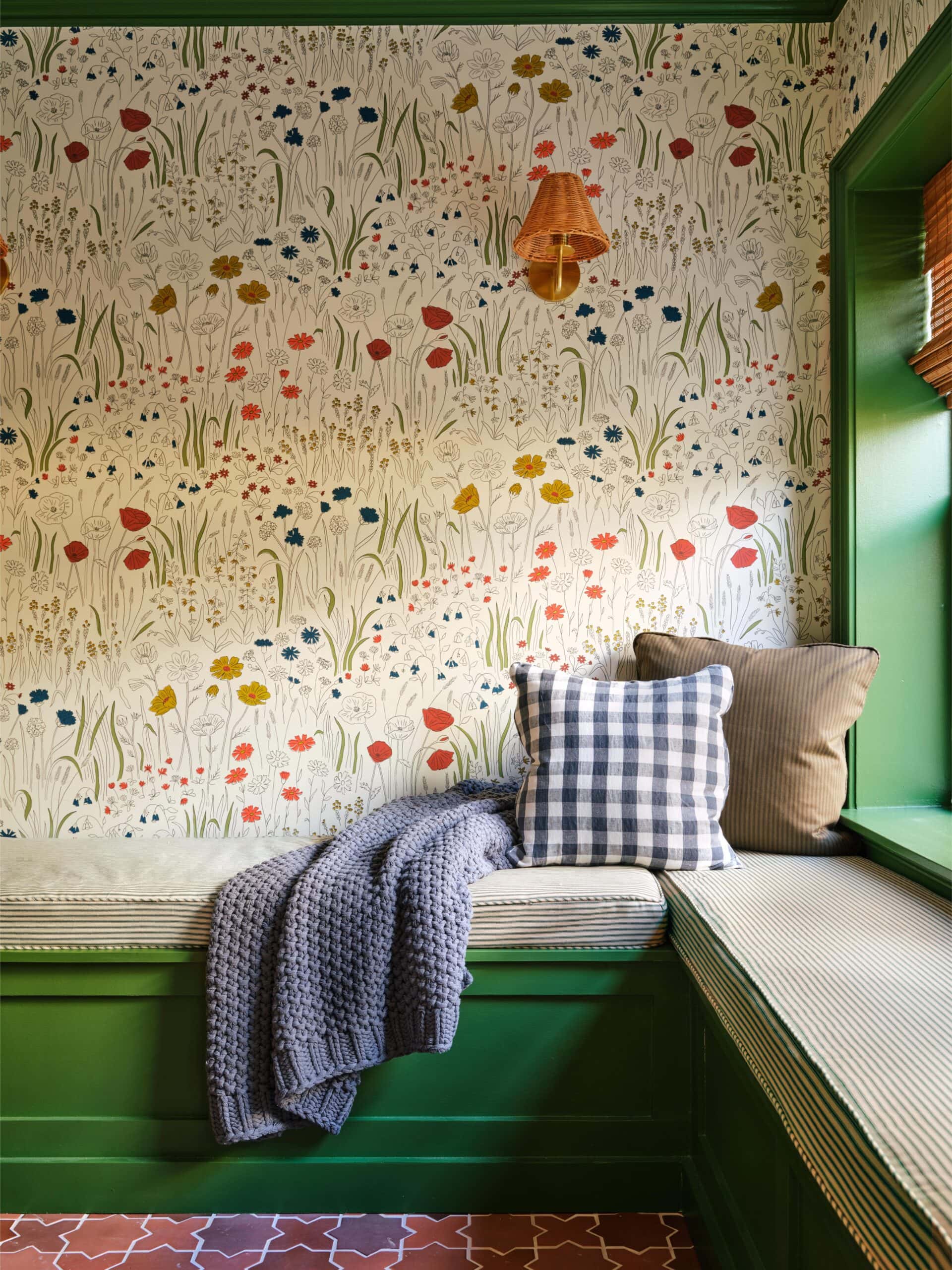 A built-in green bench with cushions and a knitted throw sits below floral wallpaper and a wall sconce in a cozy, sunlit nook.