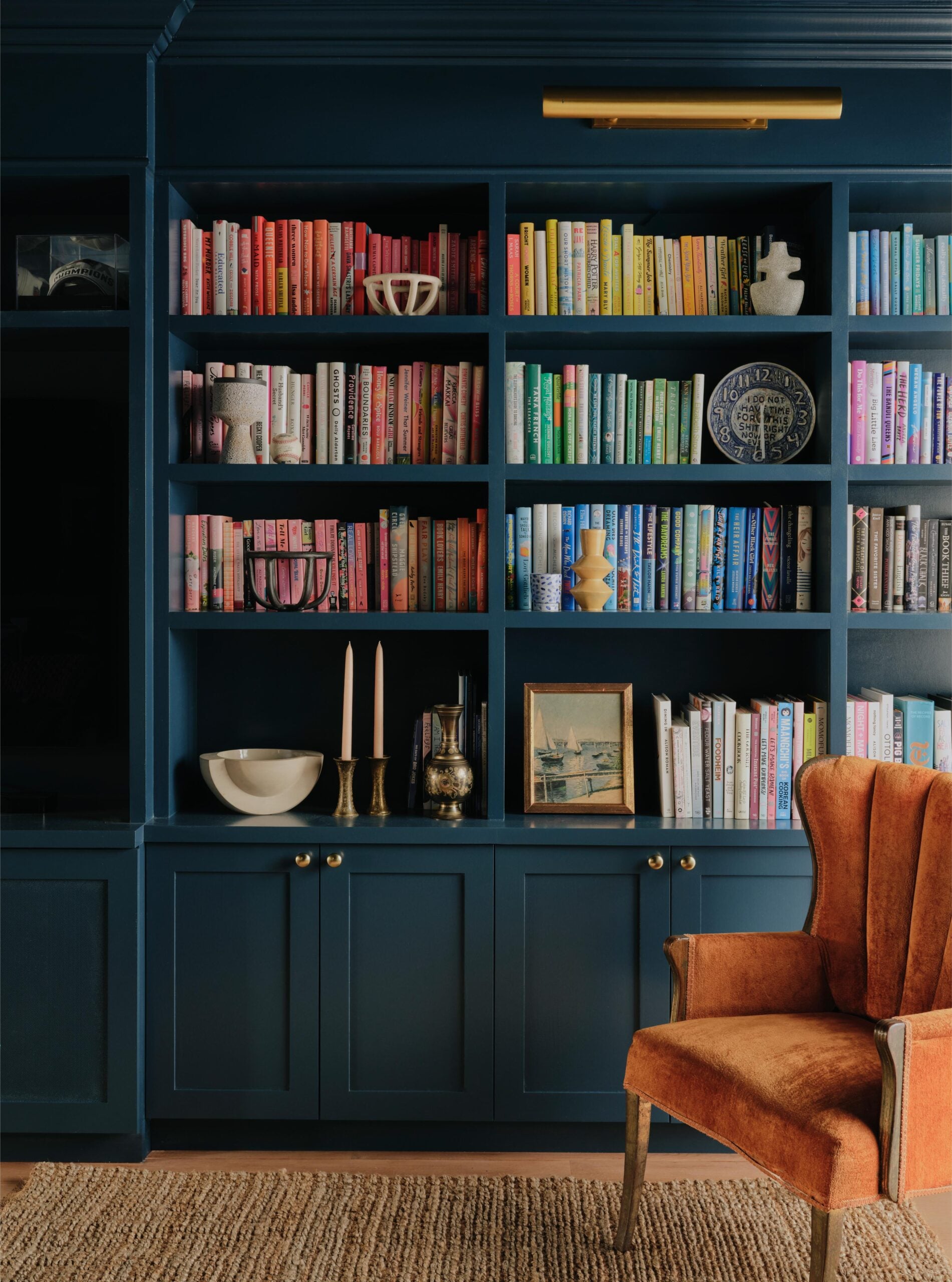 A dark blue built-in bookshelf with books arranged by color, various decorative objects, and an orange upholstered armchair on a woven rug.