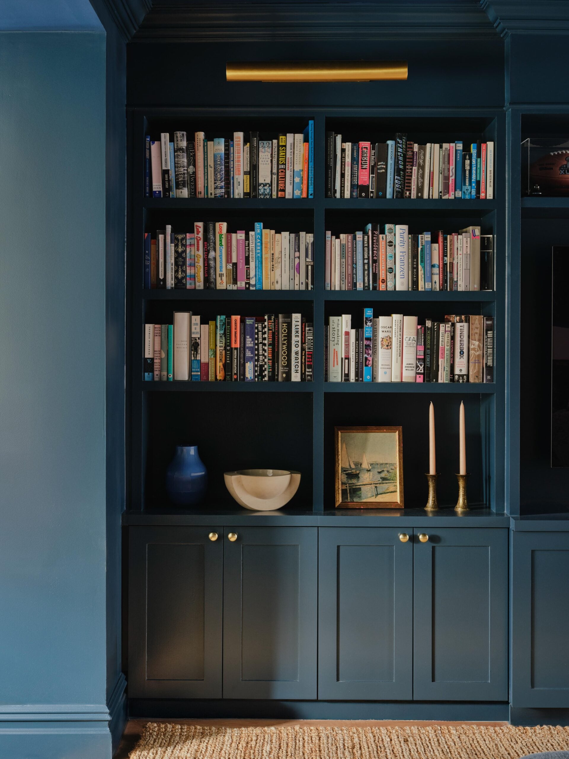 A dark blue built-in bookshelf filled with books; cabinet below holds a white bowl, blue vase, framed picture, and two pink candles in gold holders.