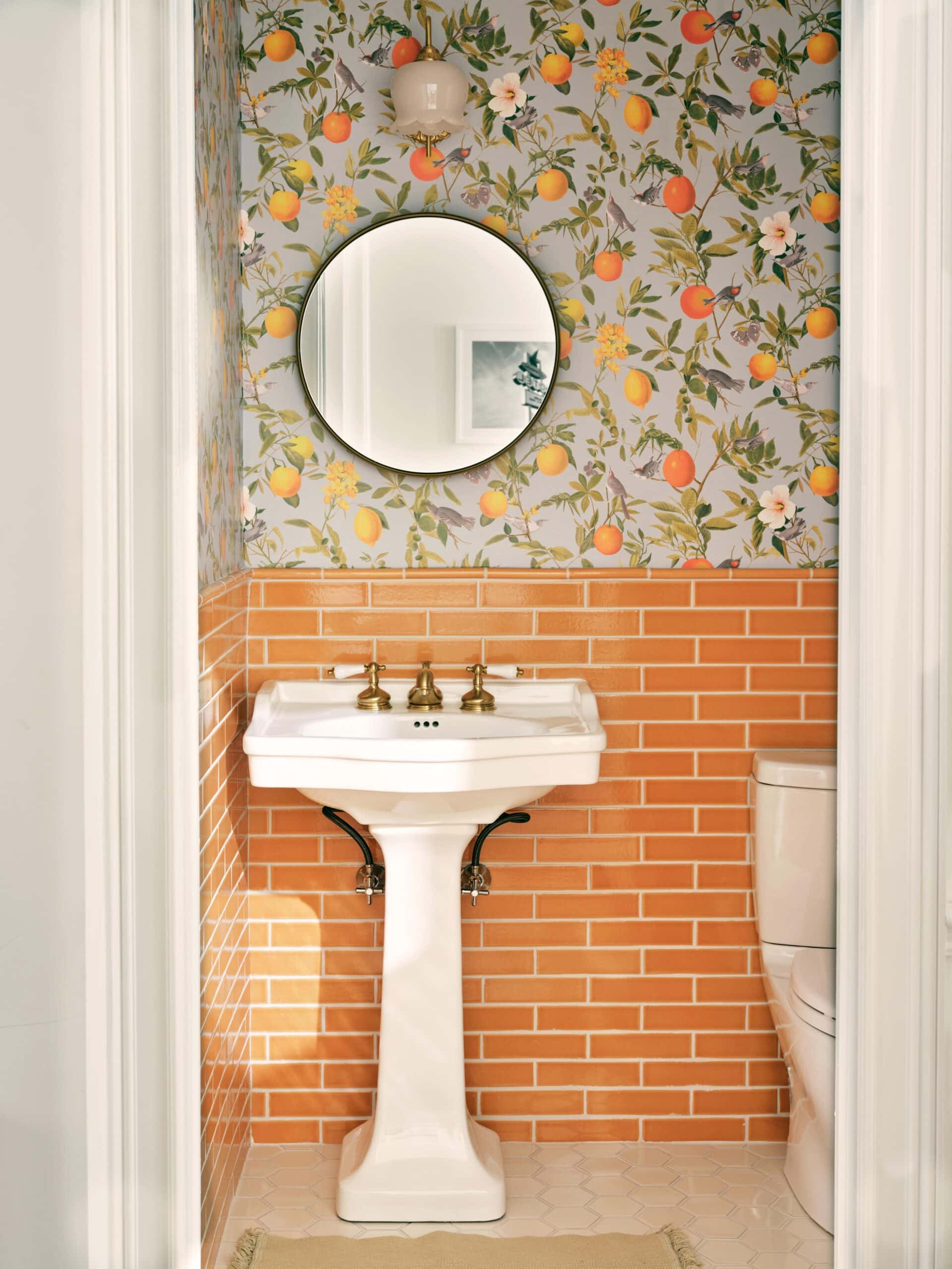 A small bathroom with citrus fruit wallpaper, orange subway tile, a pedestal sink with brass fixtures, a round mirror, and a white toilet.