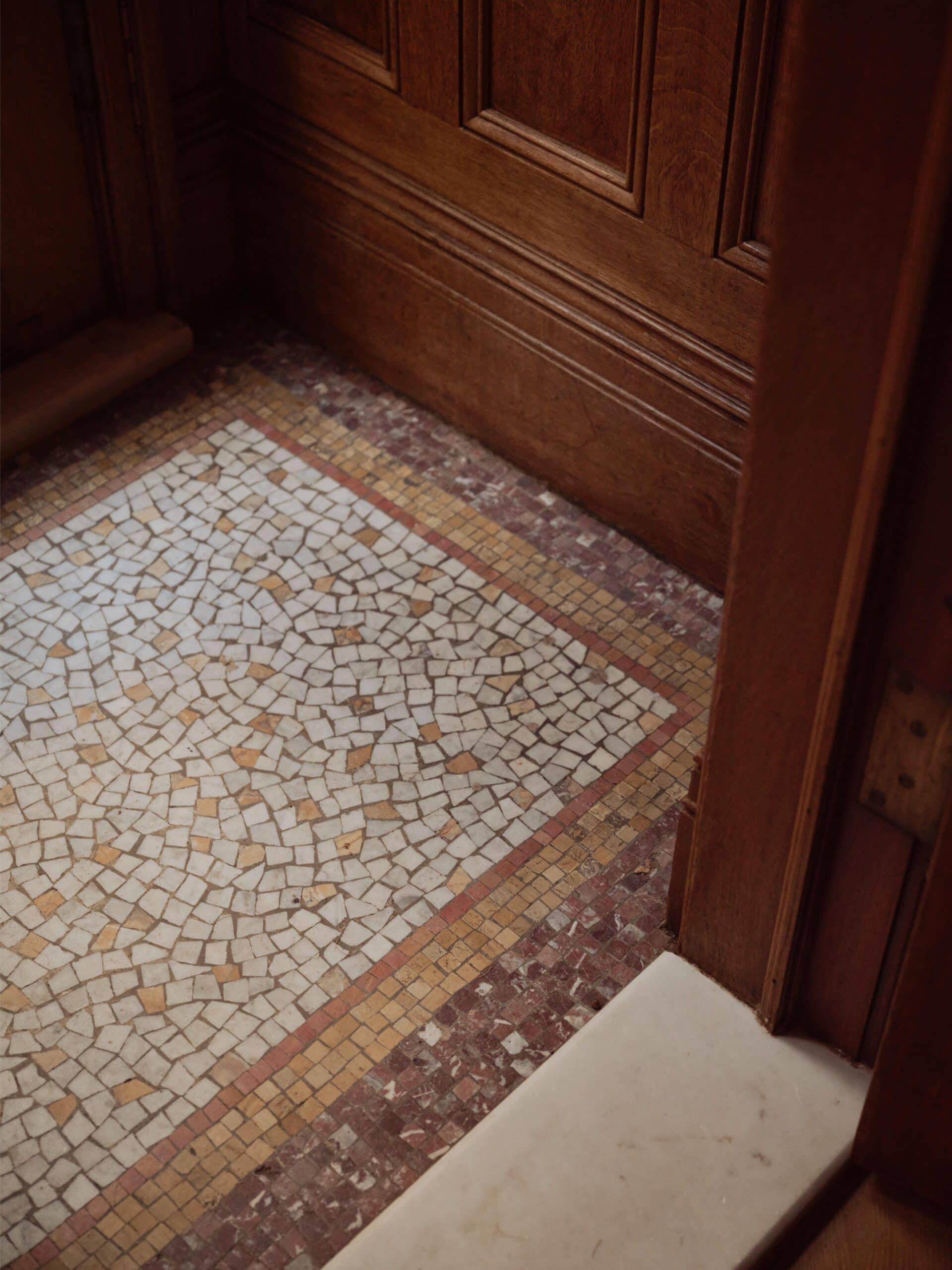 A section of mosaic tile flooring bordered by wood paneling and a stone threshold in a room corner.
