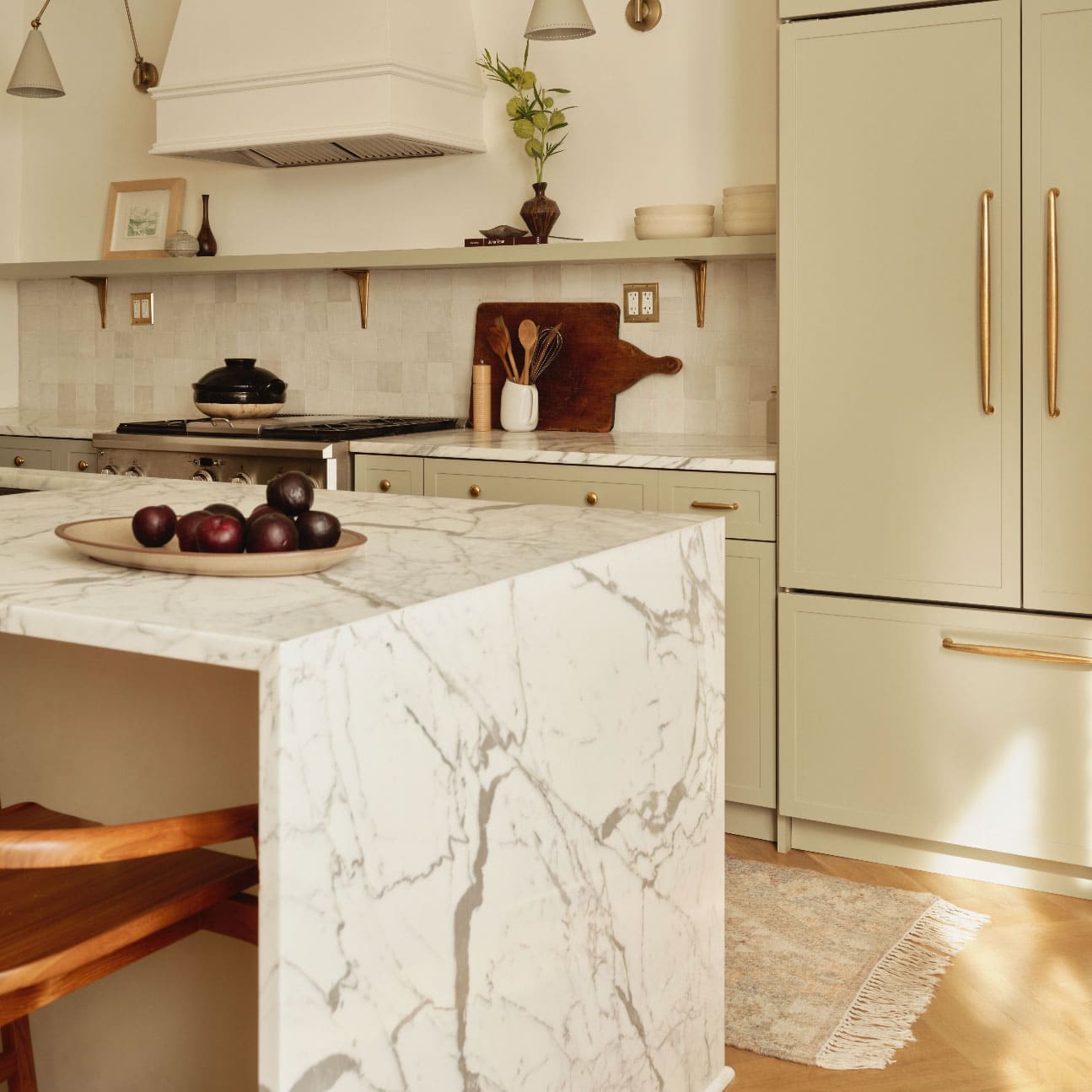Modern kitchen with light green cabinets, brass handles, marble island, wooden stools, and a bowl of plums on the counter; neutral decor and natural light.