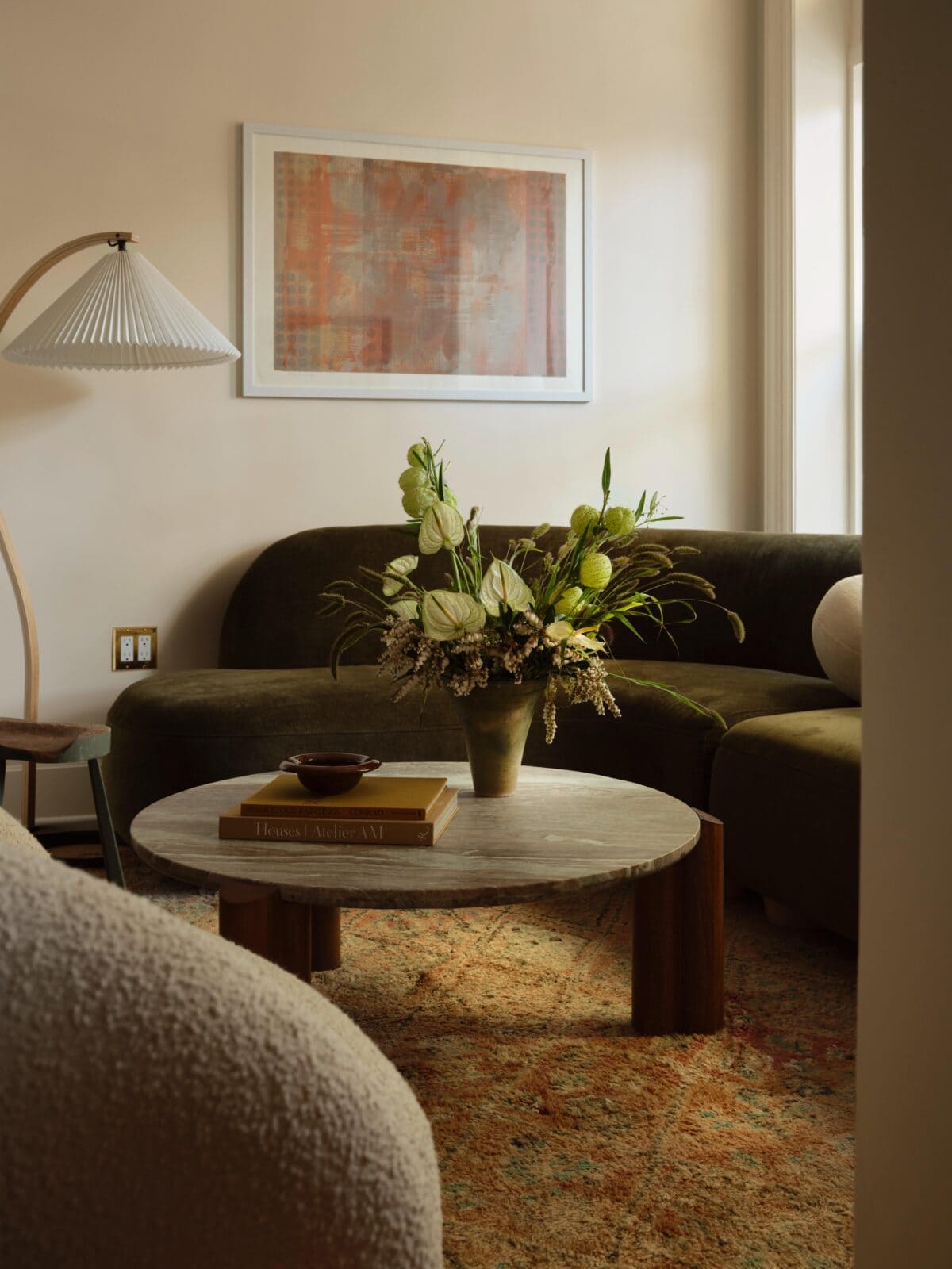 A round coffee table with a floral arrangement and books sits between curved sofas in a softly lit living room with framed art on the wall.