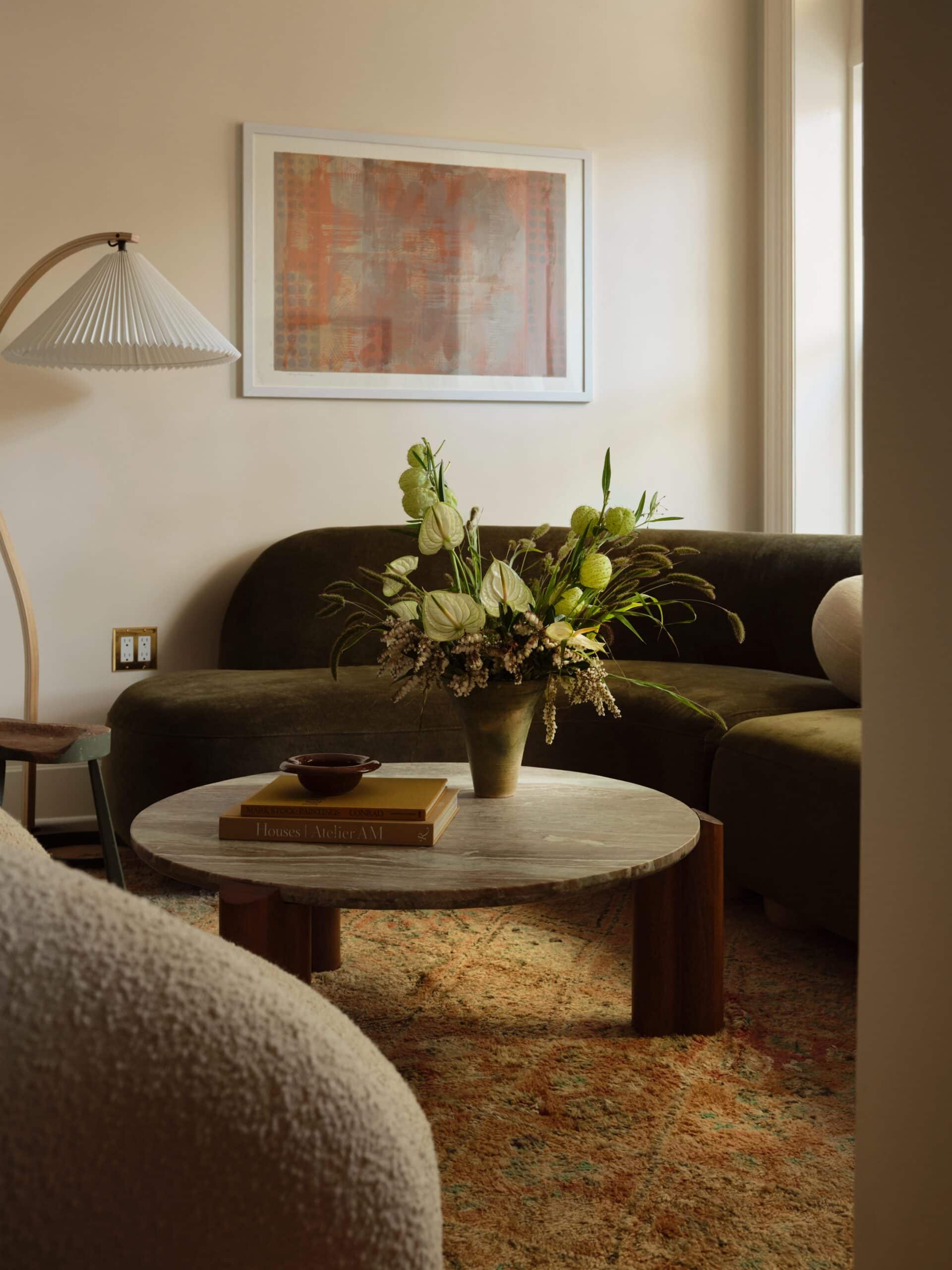 A round coffee table with a floral arrangement and books sits between curved sofas in a softly lit living room with framed art on the wall.