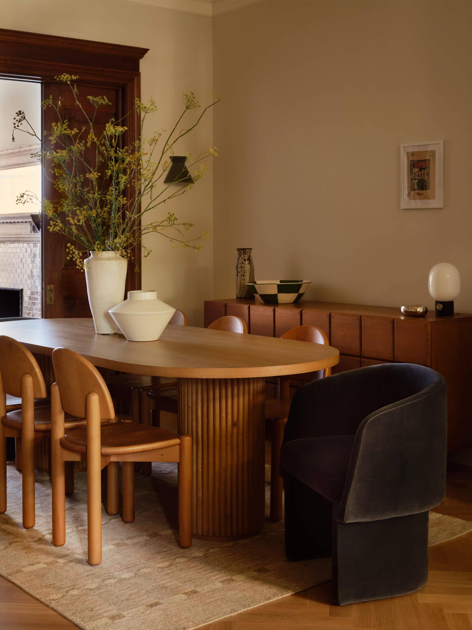Dining room with a wooden oval table, six light wood chairs, a black armchair, sideboard, decorative vases, and neutral-colored walls and decor.