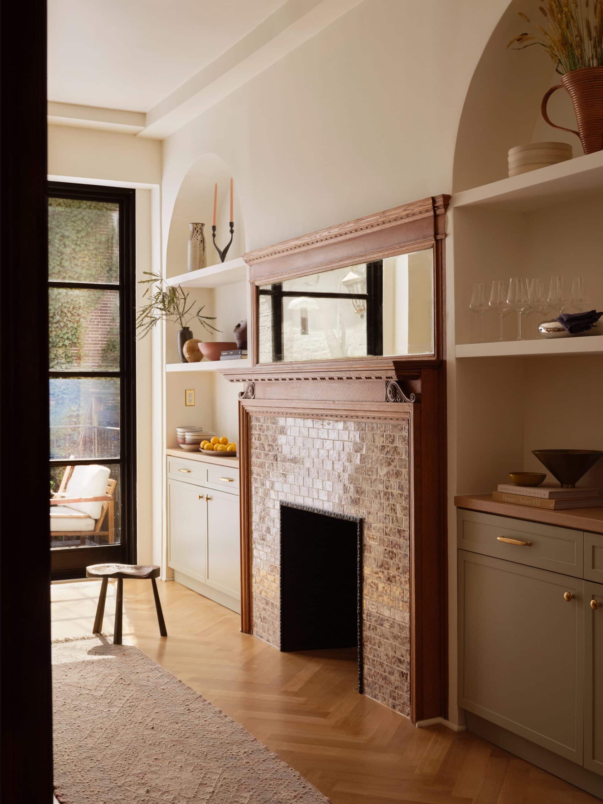 A bright kitchen features a tiled fireplace with a large mirror above, open shelves with dishes and glasses, and a windowed door leading outside.