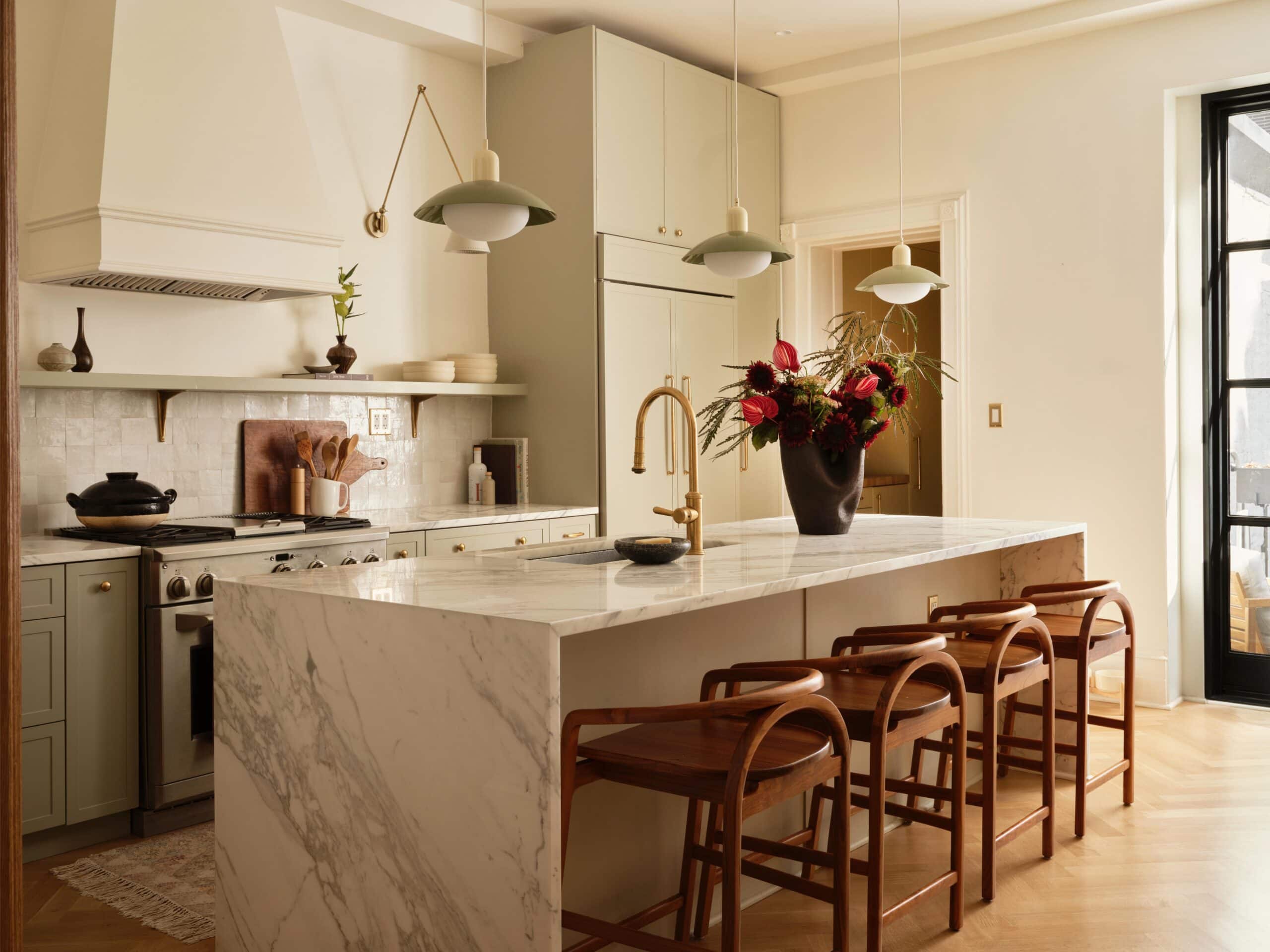A modern kitchen with a large marble island, wooden bar stools, light-colored cabinets, pendant lights, and a vase of flowers on the counter.