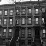 Black and white photo of a classic three-story brownstone building with stairs leading to the entrances, leafless trees in front, and all windows covered or closed.