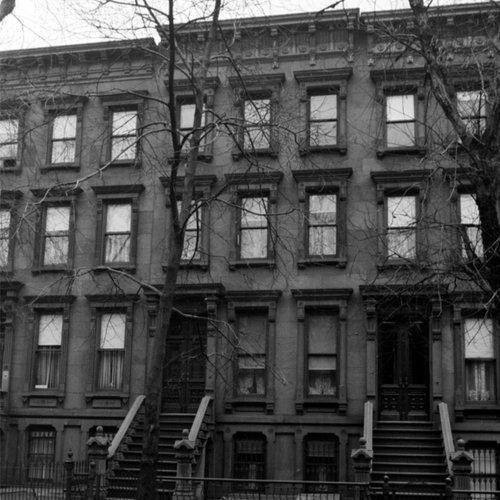Black and white photo of a classic three-story brownstone building with stairs leading to the entrances, leafless trees in front, and all windows covered or closed.