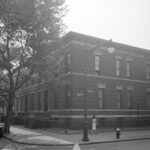 Black-and-white photo of a woman walking past a two-story brick building on a street corner, with a lamppost, tree, and fire hydrant visible.