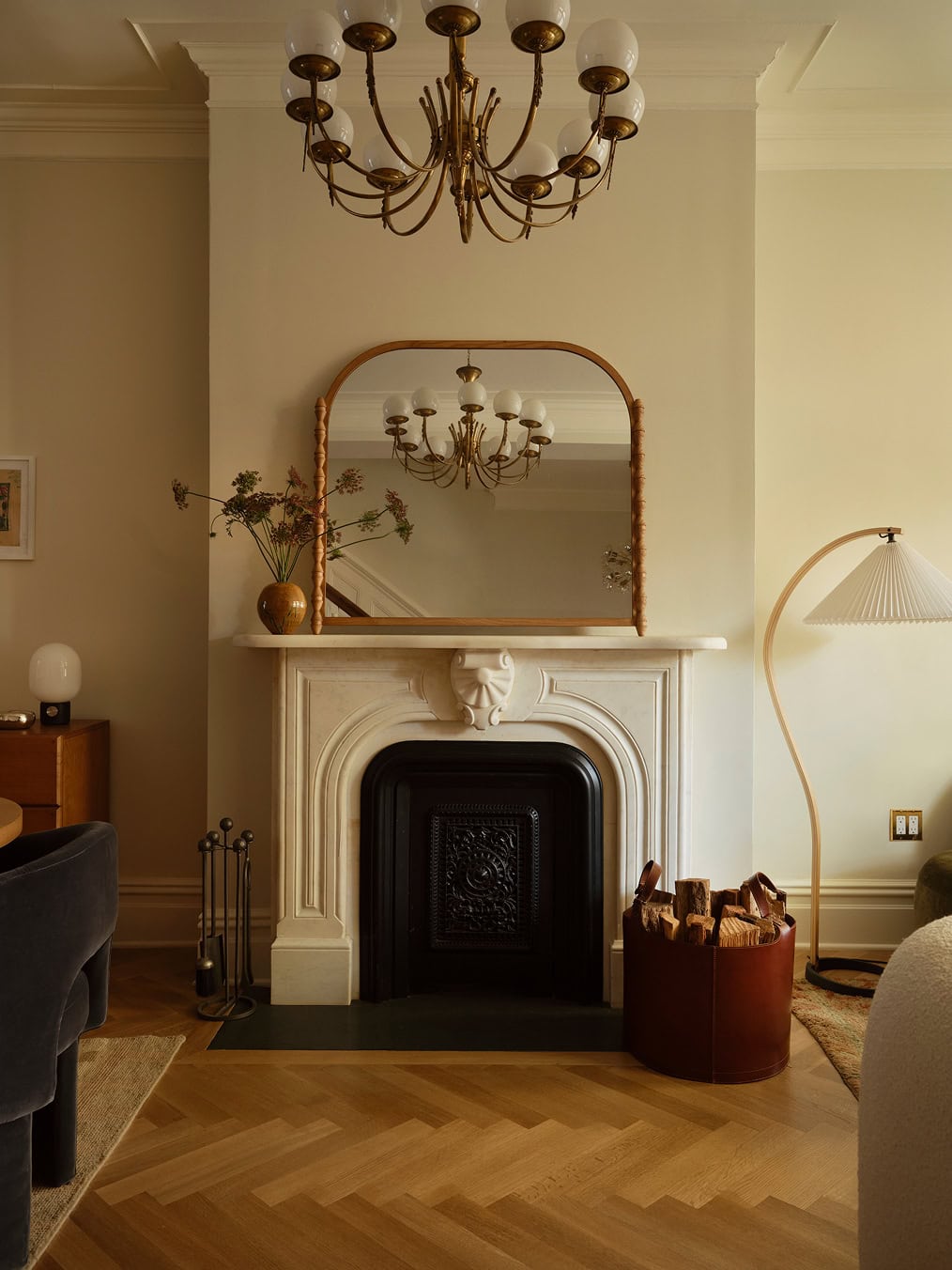 A white fireplace with a decorative mantel is topped by an arched mirror, with a brass chandelier reflected above. A floor lamp and a leather firewood holder sit nearby.