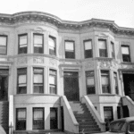 A black-and-white photo of a row of three-story brownstone townhouses with decorative facades and stoops leading to the front doors.