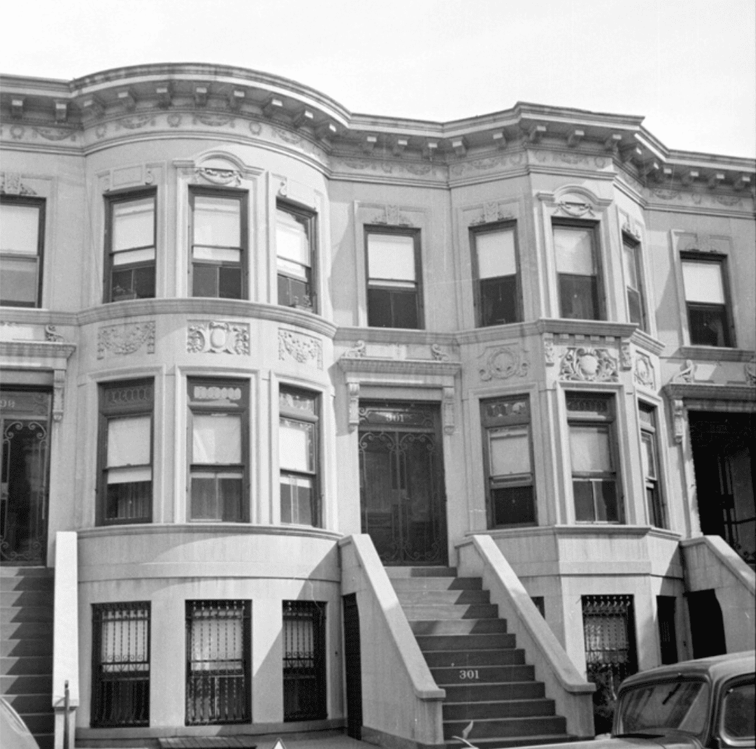 A black-and-white photo of a row of three-story brownstone townhouses with decorative facades and stoops leading to the front doors.