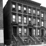 Black-and-white photo of a row of three-story brownstone buildings with stoops and metal trash cans lined up on the sidewalk in front.