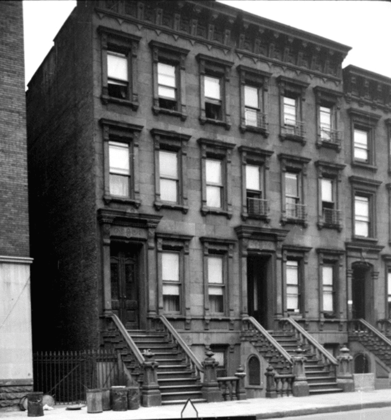 Black-and-white photo of a row of three-story brownstone buildings with stoops and metal trash cans lined up on the sidewalk in front.