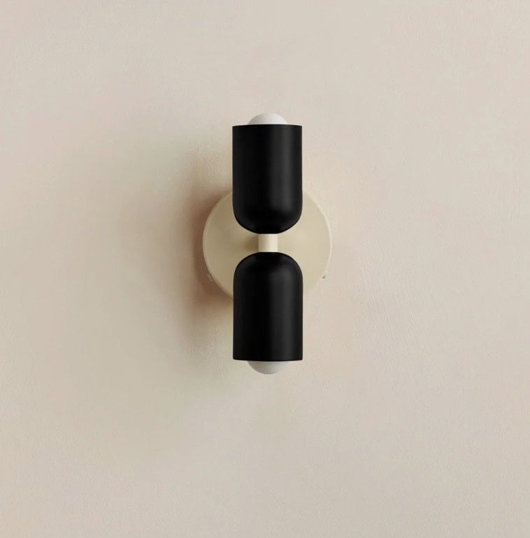 A modern wall sconce with two cylindrical black shades mounted vertically on a round beige base against a light-colored wall.