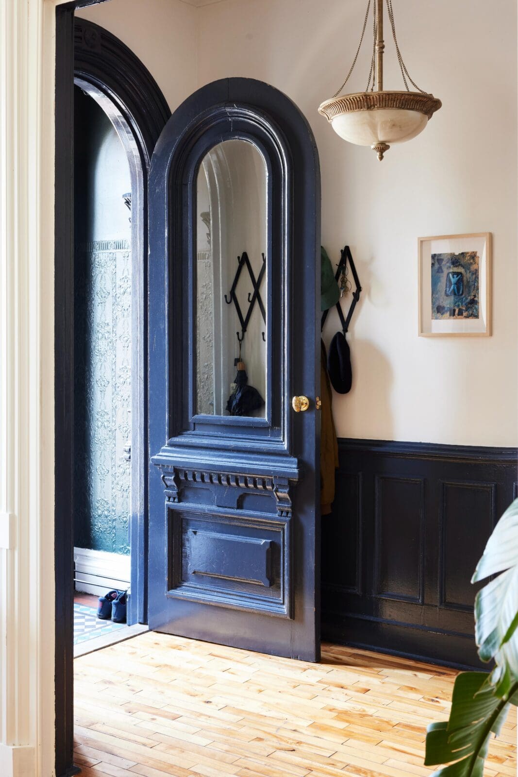 A dark blue, arched wooden door stands open, revealing a hallway with coat hooks, hats, and a framed picture on a white wall, next to a hanging light fixture.