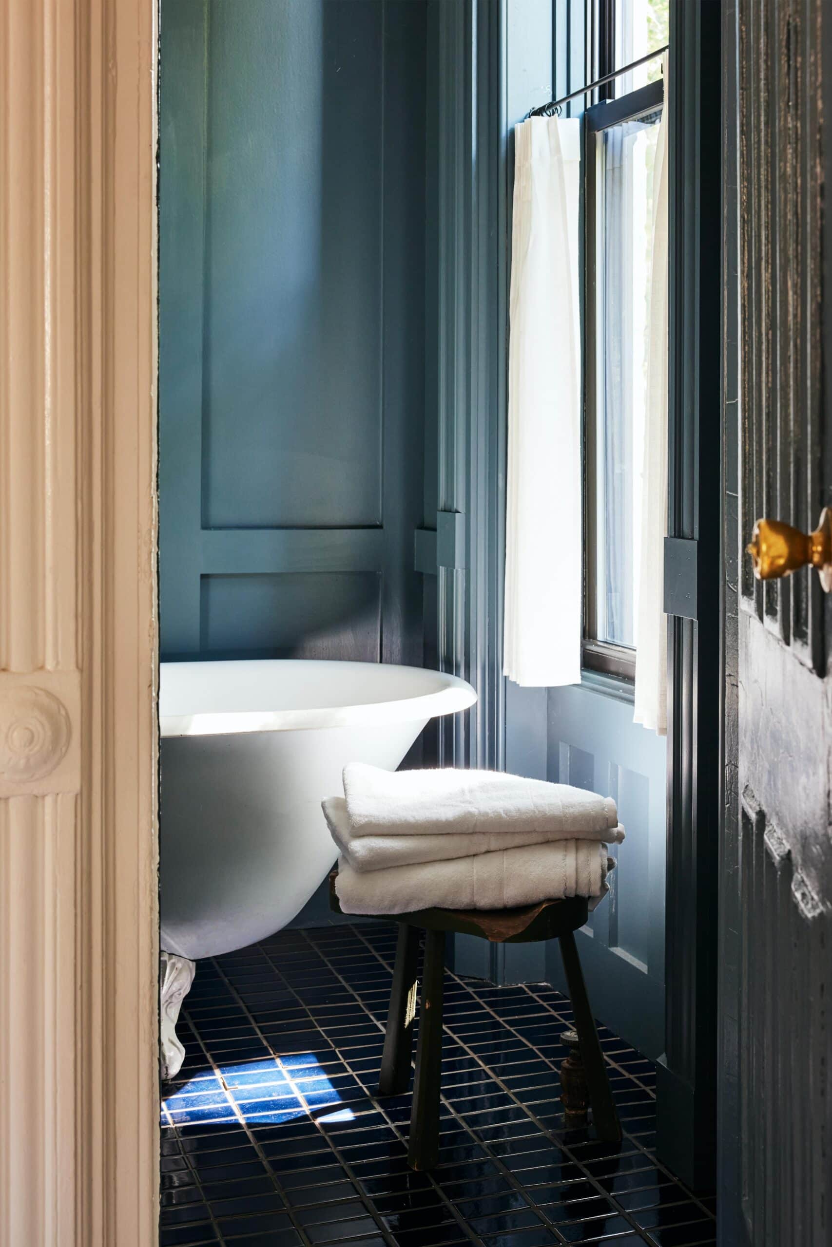 A freestanding white bathtub sits beside a window with a white curtain, and a small stool with folded towels rests on a dark tiled floor.