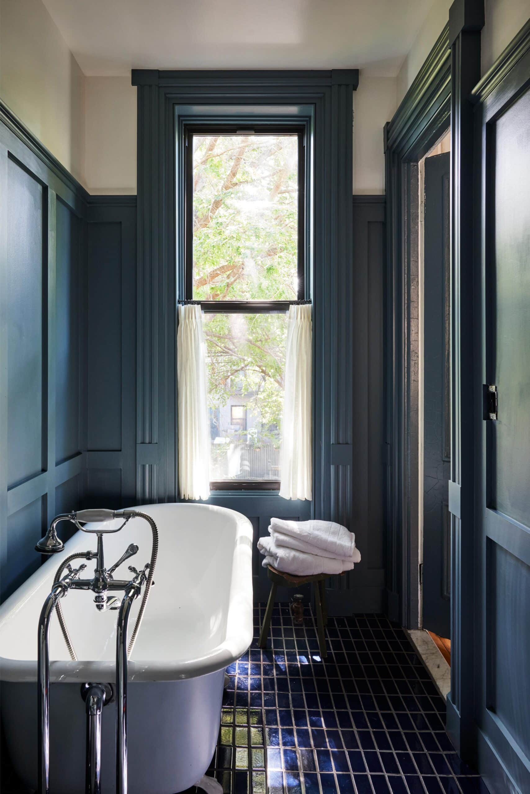 A freestanding bathtub with chrome fixtures sits next to a window with white curtains in a blue-paneled bathroom; folded towels rest on a small stool.