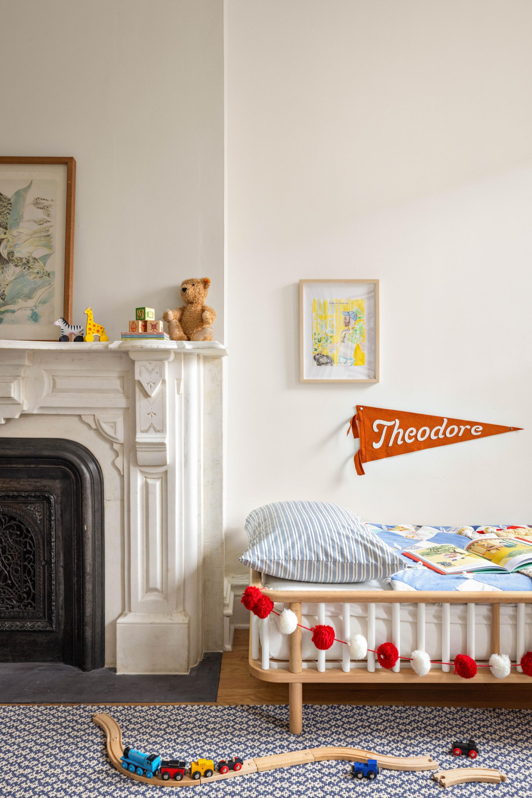 A child's bedroom with a small bed, red and white pom-pom garland, wooden toy train set, fireplace, teddy bear, and an orange pennant reading "Theodore" on the wall.