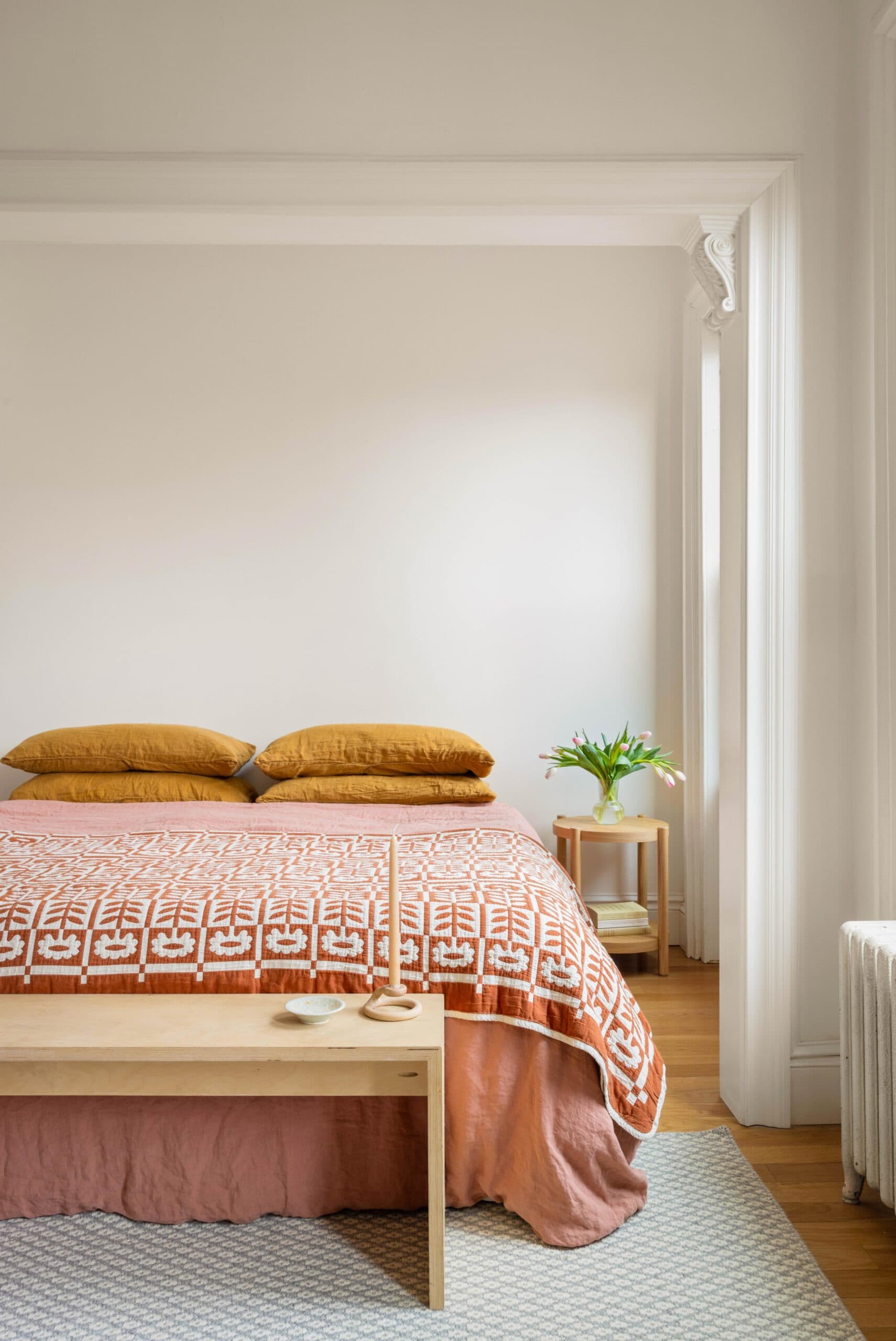 A neatly made bed with orange pillows and a patterned blanket sits in a bright room; a small nightstand with a vase of tulips is beside the bed.