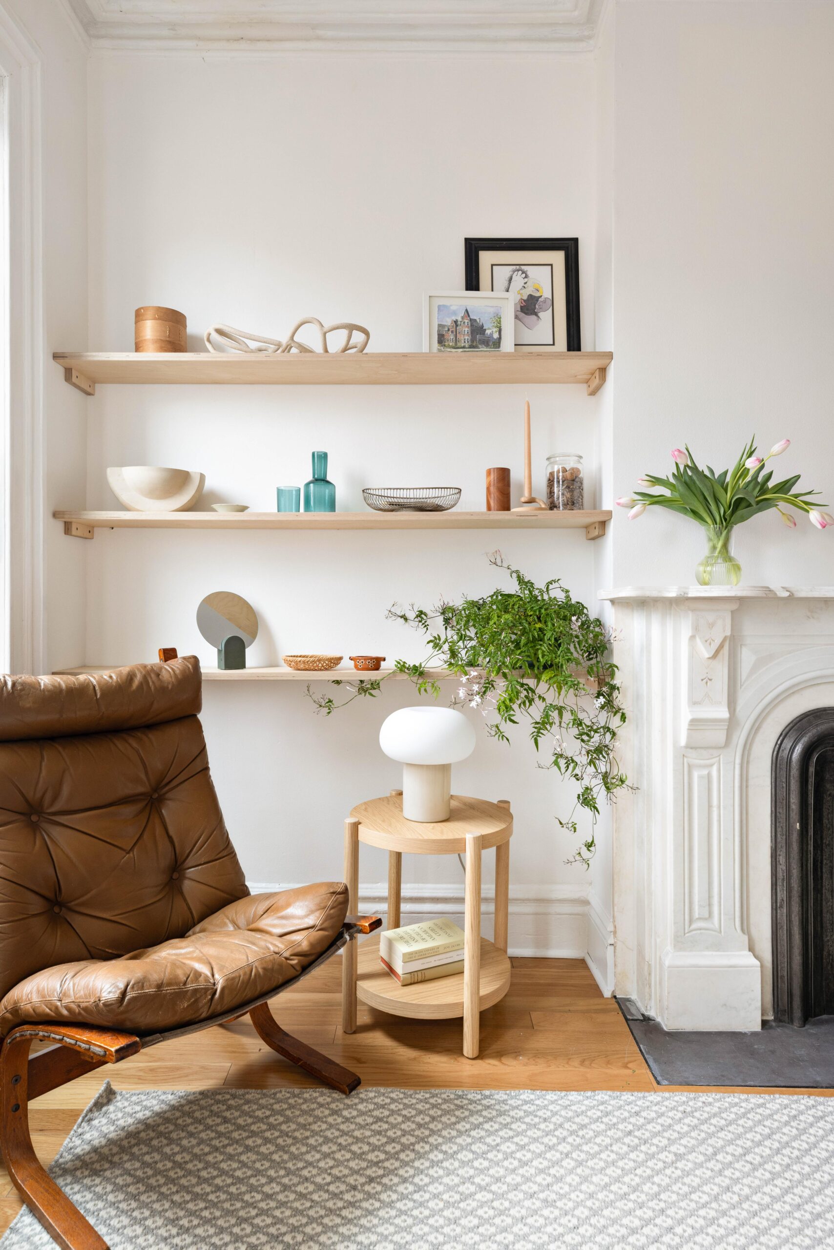 A cozy living room corner with a brown leather chair, wooden shelves holding decor items, a side table with a white lamp, and a white fireplace topped with a vase of pink tulips.