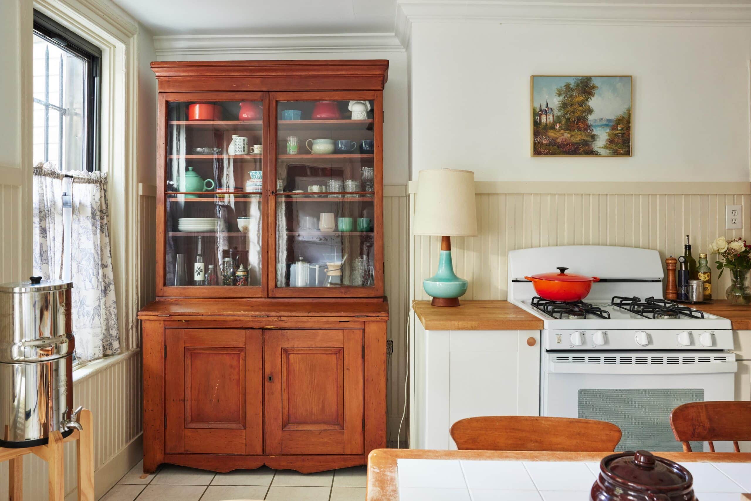 A kitchen with a wooden hutch displaying dishes, a white stove with a red pot, a table, a lamp, and a painting on the wall. Natural light comes through a window.