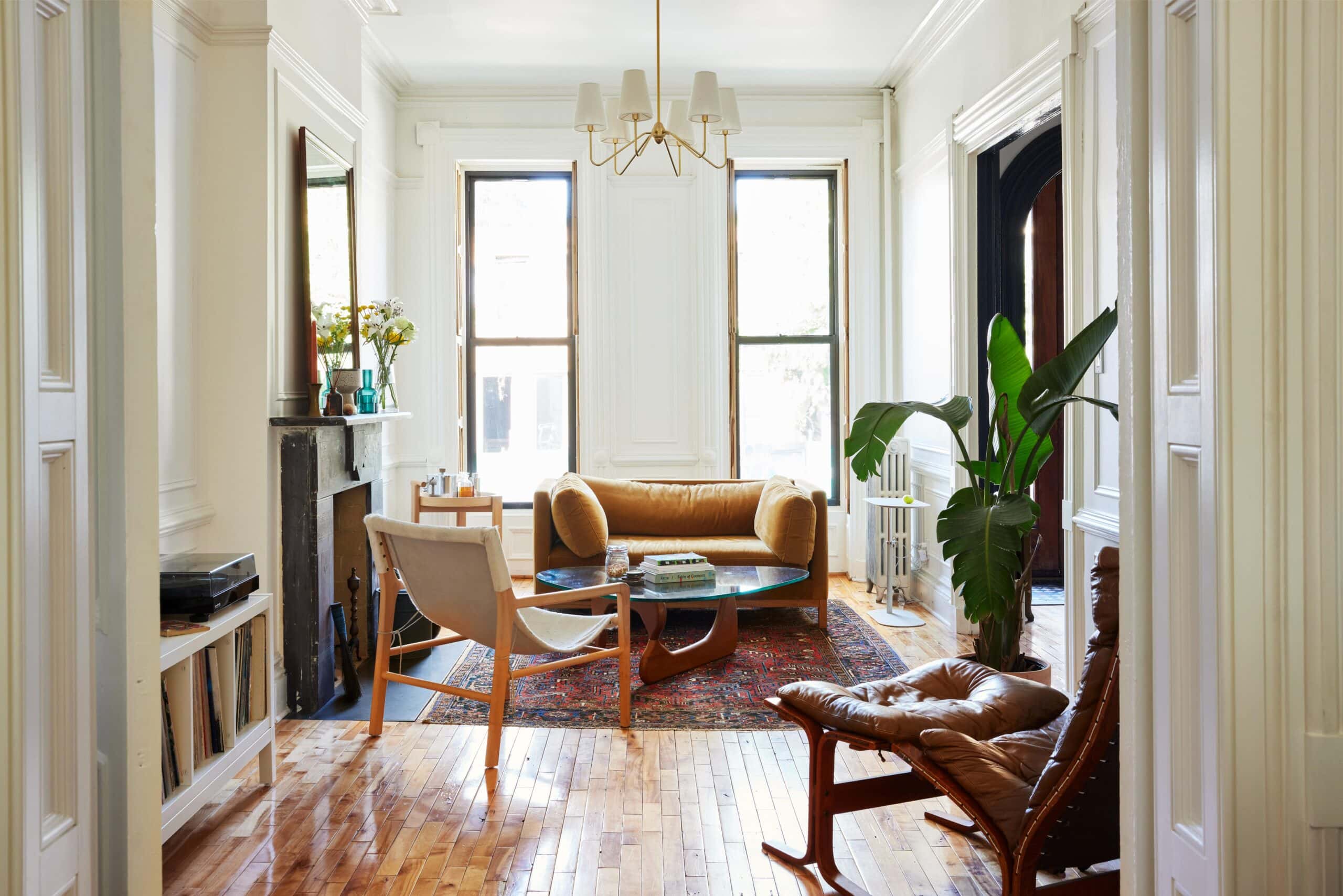 Bright living room with large windows, wooden floors, a yellow sofa, modern chairs, a coffee table, potted plant, and a patterned rug.