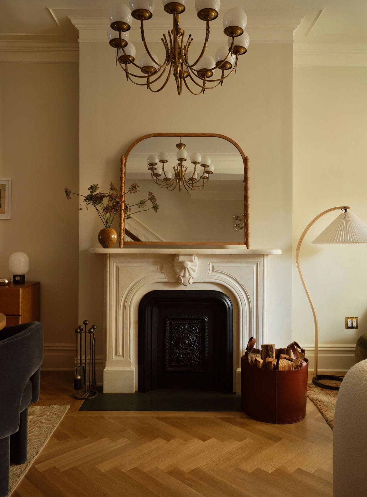 A white fireplace with an ornate mantel is topped by a large mirror. A vase with flowers, a chandelier, a floor lamp, and a leather basket with firewood decorate the room.