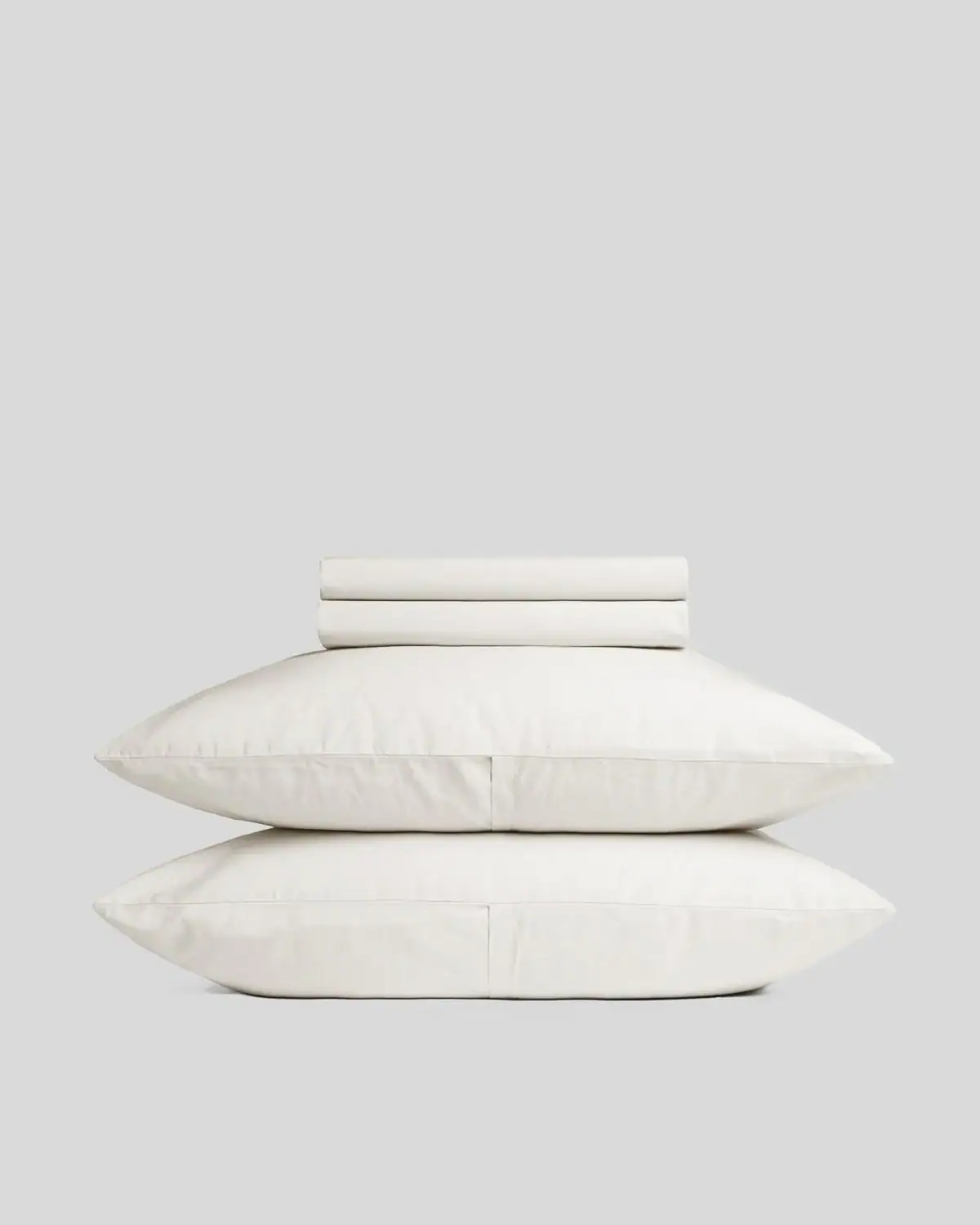 Two white pillows stacked on top of each other with a folded white sheet placed on top, set against a plain gray background.